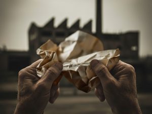 תקריב על ידיים מבוגרות, עובדות, מחזיקות מכתב דחייה קרוע למחצה מהמוסד לביטוח לאומי. ברקע, מחוץ לפוקוס, צללית של מפעל ישן. התמונה משדרת שנות עבודה, אכזבה ומאבק שקט.