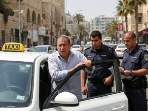 נהג מונית ליד המונית שלו שהיא מונית ישראלית לבנה עם כובע צהוב ולידו שוטרים. הנהג נראה מבולבל ומפוחד והשוטרים קשוחים